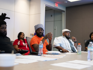 Several Black workers, women and men, sit around a conference table. In the middle of the frame, a man in an orange T-shirt is talking, looking around at the group; others are listening thoughtfully. Everyone's expression is serious.