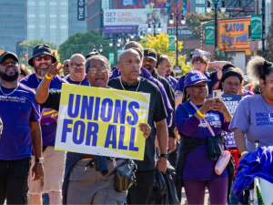 Workers march through a Detroit street, part of a Labor Day march. The people visible are mostly wearing purple SEIU T-shirts, many are Black and one holds a big printed sign readgin "Unions for all" on a yellow background. Another printed sign says "Families first, not billionaires. Not one more cut." One woman in a "Michigan home care workers" shirt pushes another in a wheelchair, also in purple gear.