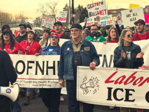People march in the street carrying various banners and signs. The most visible banners says "Labor says ICE out." Others partially visible are for two Oregon health care unions, OFNHP and ONA. Their red and green shirts (respectively) are also visible, and someone has a blue AFT shirt. Many handmade picket signs are visible, including "We are all Alex," "Unions want ICE out," "Hold ICE accountable," and "Ge(stop)o the bullshit" 