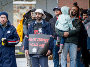 A racially diverse, young, mostly male crowd stands in front of a post office. Some people wear postal gear, others wear "Fight Like Hell" logo T-shirts, and some hold printed "First class service, first class pay" signs. One man holds a toddler in his arms. People wear coats and hats, and one open umbrella is visible in the back.