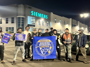 Siemens workers pose in front of the factory in the dark behind a blue banner that says Siemens Workers United.
