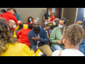 Masked people sit in small groups in a conference room, talking. They are diverse in apparent race, age, and gender.