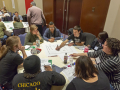 A group of 8 people sit at a round table, as one person draws a large diagram on paper on the table