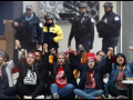 Teachers seated on sidewalk with joined hands raised; police standing behind them