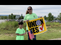 Woman holding "UNION YES" sign and child holding U.S. flag, with VW plant behind