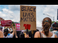Crowd viewed from back. Sign in foreground, hand-lettered: "Black Lives Matter." Signs in background, printed: "Racism is a public health crisis" with the logo of National Nurses United