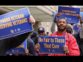 People (mainly Black men are visible) outside hold printed AFGE signs reading "Proud veterans serving veterans," "Be fair to those that care," and "VA workers deserve dignity, fairness, and respect."