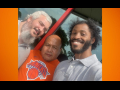 Three workers pose for a group selfie in front of the store. They read as three men of different races. The man in the middle is wearing an orange Knicks T-shirt.