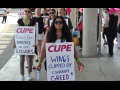 Flight attendants, mainly women, march on an airport sidewalk. Some wave pink CUPE flags and others wear sandwich-board signs with "CUPE" printed on and handwritten slogans. The two most visible say "Wings clipped by corporate greed!" and "Execs get bonuses, we get excuses." The woman in the foreground meets the camera's gaze with a serious expression; women behind her are chanting or singing.