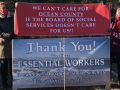 Two workers hold a banner reading “We Can't Care for Ocean County If the Board of Social Services Doesn’t Care About Us!” over the “Thank You to Our Essential Workers” sign that the Board had put up.