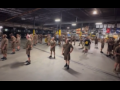 Indoors in a UPS warehouse, about 25 drivers are standing around the floor all with their backs to a manager with a yellow clipboard