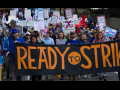Big crowd marches behind a giant black banner hand-painted in orange "Ready to Strike." Many people hold handmade signs on sticks. A person near the front with long pink hair and reflecting sunglasses looks defiantly into the camera. A person in a blue beanie punches a tattooed arm into the air, raising a fist. Farther back, glimpse someone in a red inflatable lobster suit.