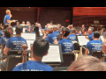 Orchestra musicians on a stage, apparently performing or rehearsing (conductor's baton is raised). Everyone, including the conductor, is wearing matching blue T-shirts that say "Philadelphia Musicians Union Local 77-274" on the back.