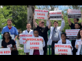 A diverse group of people, mostly in white lab coats, stand outside holding printed signs that say, in red on white, "Medicaid saves lives." Other signs say "Code red: patients in crisis" and "Our patients rely on Medicaid to get the care they need. This bill would end that."