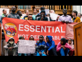 A diverse and serious crowd of people stands close together inside what looks like a church, in front of organ pipes, holding a big printed red banner that says "Essential not deportable" with the logo of Escucha Mi Voz. The person in the middle is speaking into a mic. Several kids sit in front and one holds a hand-lettered sign: "Free my grandma! Free her! I hate ICE!"