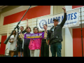 Six workers, of various races and genders, stand together on a ballroom stage with a big "Labor Notes" banner hanging behind them. Three are raising fists in the air, and one holds out a purple "SEIU Local 26, #UnionsForAll" scarf.