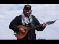 Emmett Doyle, a bearded white man wearing a "Class war veteran" hat, a keffiyeh, and a Carhartt jacket, plays an Irish bouzouki. He is outdoors, silhouetted against a blue sky with thin white clouds.