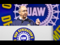 Shawn Fain, a white man with glasses, speaks at a podium. Behind him (and on the podium) is the UAW logo in white on a blue background. Yellow letters around it say (partly obscured) "Our Generation's Defining Moment."