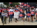 A group of workers pose for the camera on a city sidewalk. They are diverse in race, gender, and age. Many hold printed red and blue signs that say "PECO workers on strike!" One holds a big hand-drawn sign: "26+666+614=1. IBEW united!"