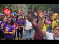 A large crowd of people, mostly Black women, rallies in Manhattan's Foley Square. Many wear purple 1199 T-shirts. Some hold yellow 1199 signs, or red ones shaped like hearts with the message "We are the heart of New York healthcare." The mood is active, with many arms reaching up into the air and mouths opening to speak, chant, or sing.