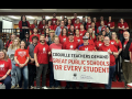 A group of dozens of teachers wearing red shirts gather behind a union banner.
