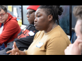 A Black woman in a tan shirt speaks while three men, white and Black, look on.