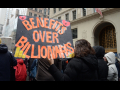 Marchers walk away from the camera with a giant orange on black sign, “Benefits over Billionaires”