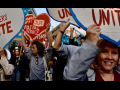 Protesters crowd towards the camera with heart-shaped signs saying “workers unite” and “hands off our postal service”