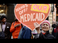 An older person holds up a handmade orange sign saying “Hands off our Medicaid”