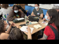 A group of masked people sit around a table discussing something intensely.