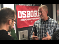 A man in a checkered shirt talks to two people with their backs to the camera in front of a red “Dan Osborne Independent for Senate” banner.