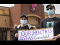 Two taco bell workers with masks holding up a sign in front of a taco bell that says "our health is just as essential."