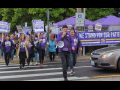 Workers in purple SEIU Local 49 T-shirts march across a street. Some carry picket signs that say SEIU and some other words too small to read. A large purple banner says "We stand for our patients." The person in front is talking into a bullhorn and others appear to be chanting. 