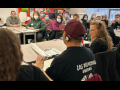 People sit in a classroom. Most are looking attentively at a person in the foreground who must be speaking, but has his back to us. He is wearing an interpretation headset and the back of his T-shirt says "Los Deliveristas Unidos," Spanish for united delivery workers.