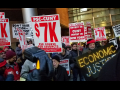 crowd holding signs including "PSC-CUNY $7k per course for adjuncts," "invest in CUNY, invest in New York," and "ECONOMIC JUSTICE" spelled out in lights
