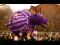 A crowd surrounds a large festive purple piggy-bank piñata suspended above them. It is labeled NYU and is in school colors, purple and gold.