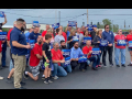 A group of workers in red, blue, or grey shirts hold signs saying “United for a Strong Contract,” and “COLA and Fair Pay Now”  