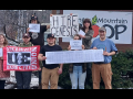 Eight people outside face the camera with signs, including “Hire Genesis” and a UE banner. 