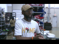 A Black man in a baseball cap and a T-shirt that says "Home" with the shape of Africa for the "O" speaks to the camera. He is standing in a warehouse, in front of shelves stacked high with folded apparel . Another Black man can be seen behind him, working.