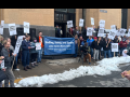 Postal workers rally outside a post office. There is some snow on the ground. A big blue banner says "Staffing, Safety, and Service: Letter Carriers Need a Raise! NALC Branch 9, Minneapolis." Printed picket signs say "End Mandatory Overtime," "First Class Service, First Class Pay," and "Fair Contract Now."