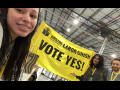 Inside a warehouse three young people smile as they unveil a bright yellow banner that says "Amazon Labor Union, Vote Yes"