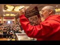Seen from the side/back, Shawn Fain in a red hoodie holds up a battered purple copy of A Troublemaker's Handbook. Arrayed in front of him can be seen a huge seated crowd in the ballroom for the closing plenary.