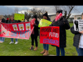 People stand on grass with signs. A red banner says "Seven months without a contract, OMG!" with stylized images of Edward Munch's painting "The Scream." Another sign says "More sick leave" in English, Spanish, and Russian. Another says, in Spanish,"Provide money for the future of students." Another says "Woodburn teachers don't want to strike, but we will." 