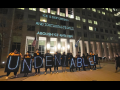 People holding lit signs spelling out "UNDENIABLE" stand in front of a building. On the building are projected the words: "ICE IS DEPORTING AND TORTURING PEOPLE! ABOLISH ICE AND CBP!"