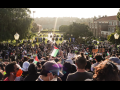 A large crowd, mostly viewed from behind, is arrayed on a hillside or steps of a university campus; banners identify it as UCLA. A few in the crowd hold Palestinian flags and one handmade sign reads "Stay strong." A row of parked vehicles is visible in the distance, at the foot of the hill, backs to the crowd. Dramatic sunlight silhouettes everyone.