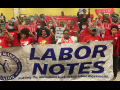 People stand indoors behind a big Labor Notes banner. Most are wearing red shirts and have their fists in the air. Several farther back are holding up smaller red "Organize the South" banners.