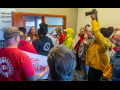At least a dozen activists crowd up to the door of a VU Manufacturing office, where a company flack is gesuring widely and trying to fend them off. One activist is holding a camera aloft, others holding signs or wearing UAW or Labor Notes gear. A woman 