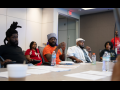 Several Black workers, women and men, sit around a conference table. In the middle of the frame, a man in an orange T-shirt is talking, looking around at the group; others are listening thoughtfully. Everyone's expression is serious.