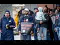 A racially diverse, young, mostly male crowd stands in front of a post office. Some people wear postal gear, others wear "Fight Like Hell" logo T-shirts, and some hold printed "First class service, first class pay" signs. One man holds a toddler in his arms. People wear coats and hats, and one open umbrella is visible in the back.