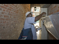 A letter carrier with a big shoulder bag puts mail in a grouped mailbox in an alley, he is backlit.